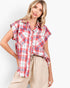 A woman modeling a red, white, and blue plaid short-sleeve button-up shirt tucked into beige drawstring pants against a light gray studio background.