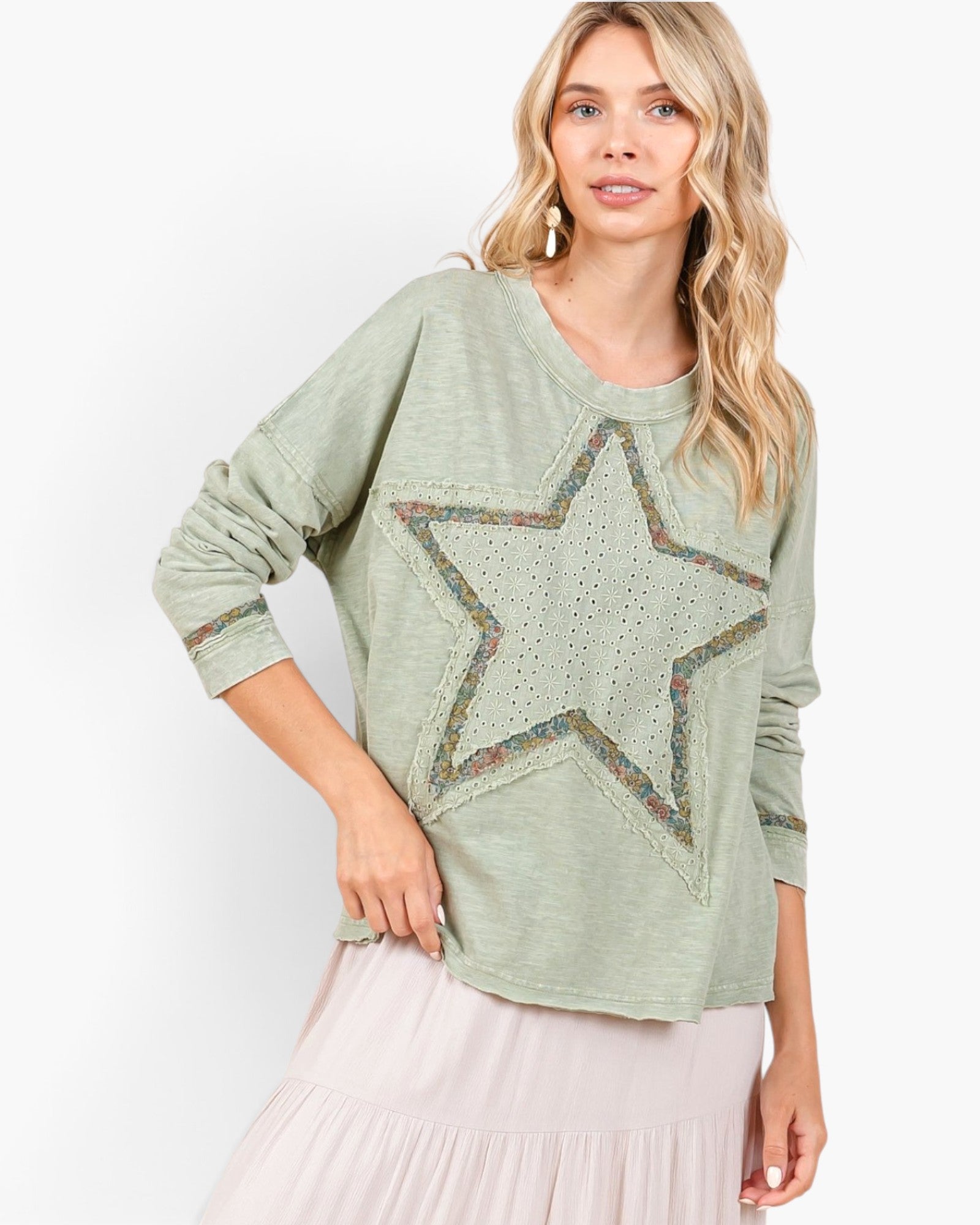 A woman wearing a sage green long-sleeve top with a large embroidered star on the front, paired with a light pink tiered skirt, against a plain white background.