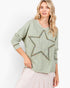 A woman wearing a sage green long-sleeve top with a large embroidered star on the front, paired with a light pink tiered skirt, against a plain white background.