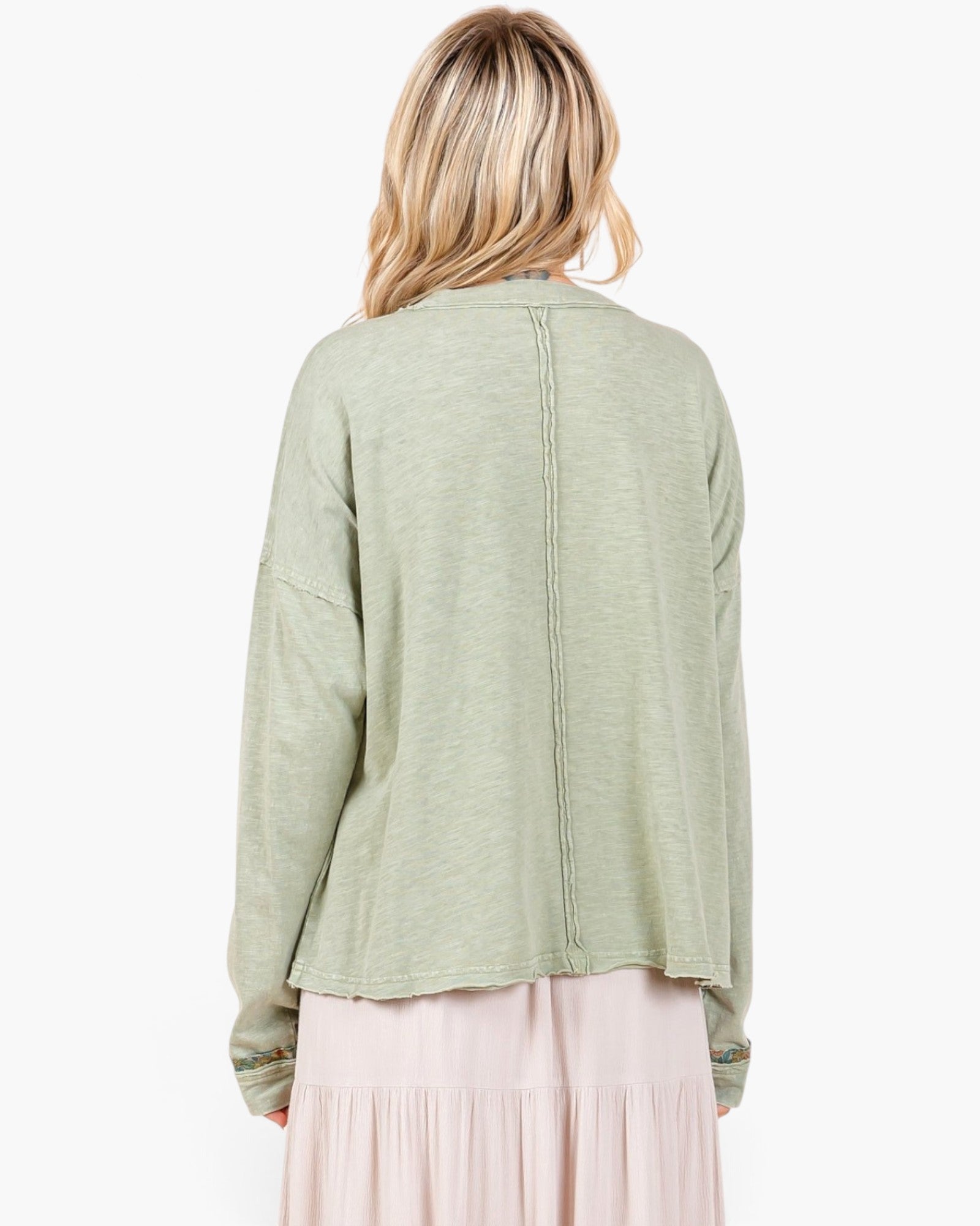 A woman viewed from the back wearing a light sage green long-sleeve top and a light pink skirt against a plain white background.