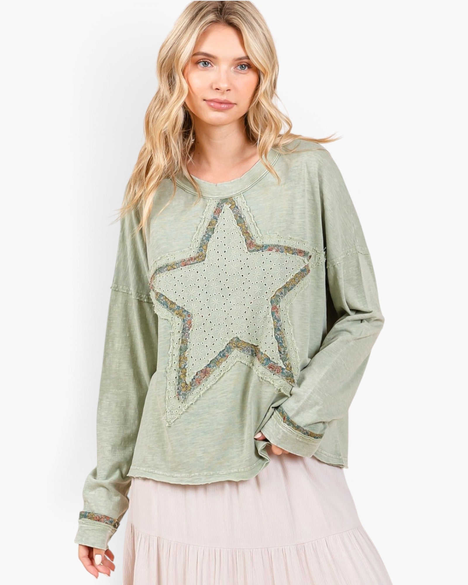 A woman wearing a sage green long-sleeve top with a large embroidered star on the front, paired with a light pink skirt, against a plain white background.