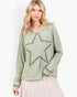 A woman wearing a sage green long-sleeve top with a large embroidered star on the front, paired with a light pink skirt, against a plain white background.