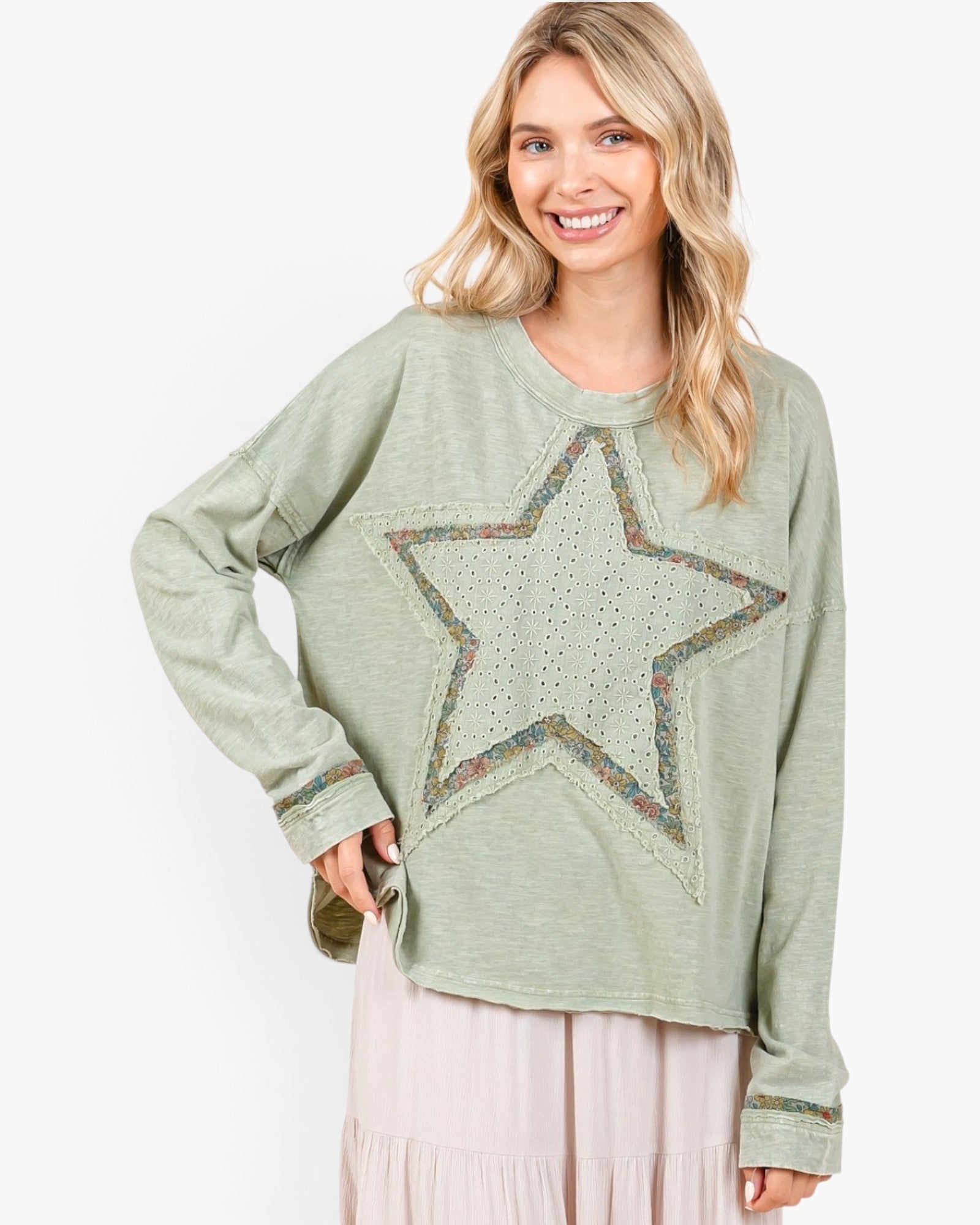 A woman wearing a sage green long-sleeve top with a large star-shaped embroidered patch on the front, paired with a light pink skirt, against a plain white background.