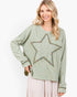 A woman wearing a sage green long-sleeve top with a large star-shaped embroidered patch on the front, paired with a light pink skirt, against a plain white background.