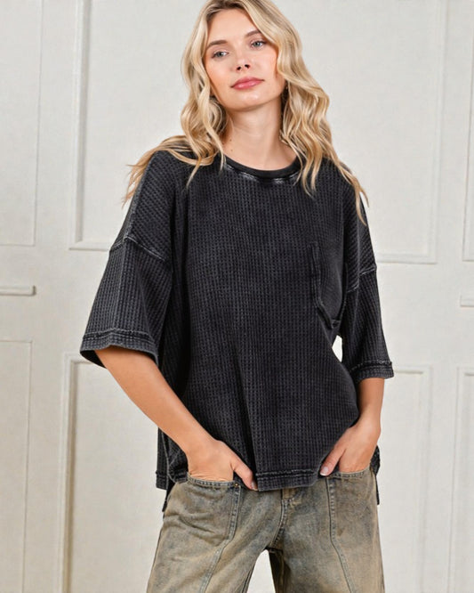 Woman wearing a black textured shirt and beige pants against a white paneled wall.