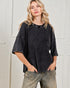 Woman wearing a black textured shirt and beige pants against a white paneled wall.