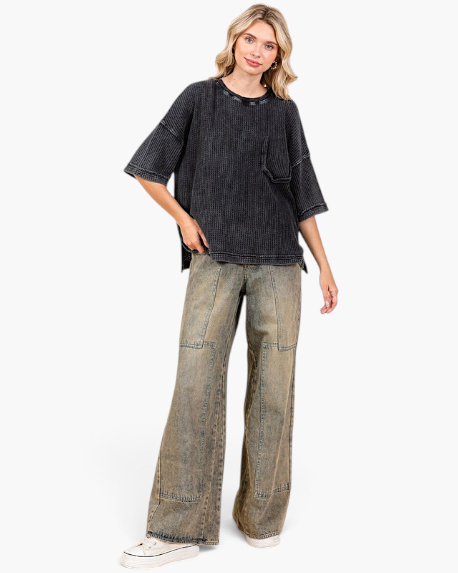 A woman modeling an oversized faded black short-sleeve top and wide-leg acid-wash jeans with white sneakers, photographed against a clean white studio background.