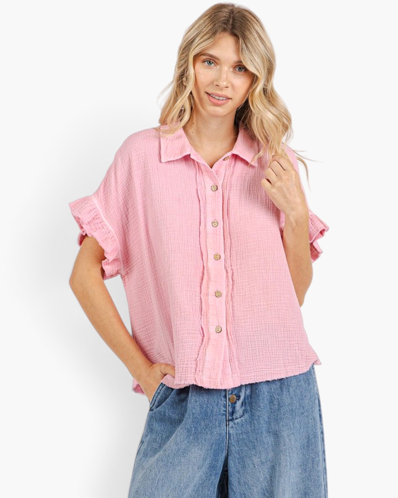 A woman modeling a light pink textured short-sleeve button-up shirt with ruffled cuffs, paired with blue jeans, against a plain white studio background.