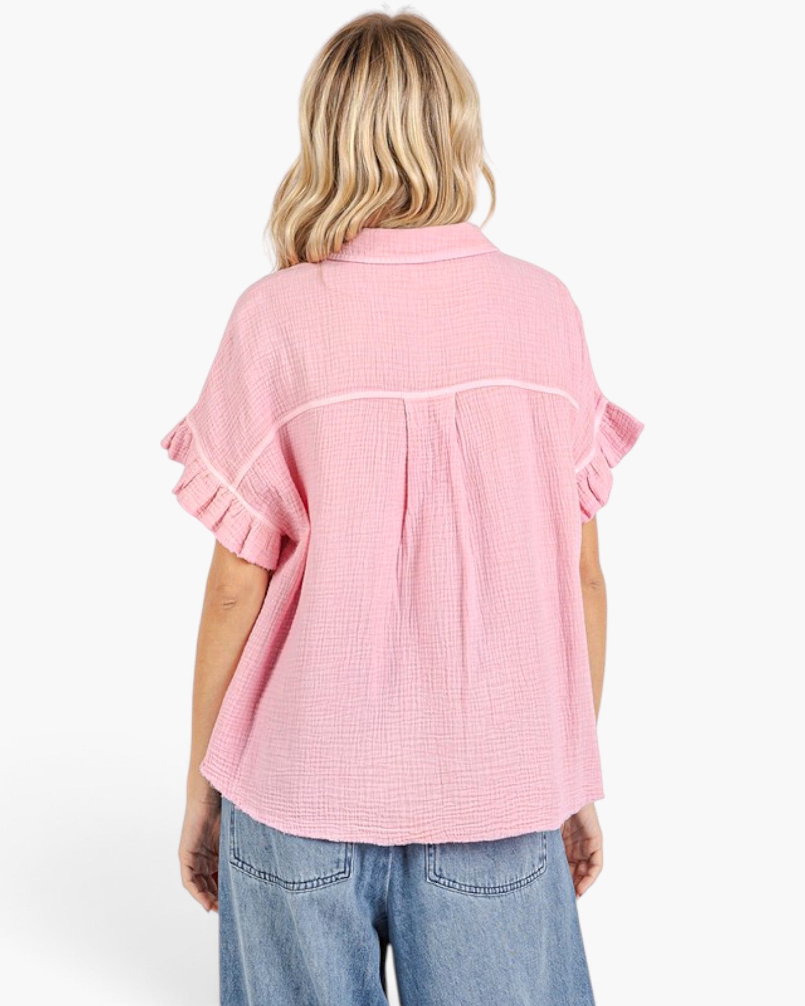 A woman viewed from the back wearing a light pink textured short-sleeve blouse with ruffled cuffs and a pleated back detail, paired with blue jeans against a plain white background.