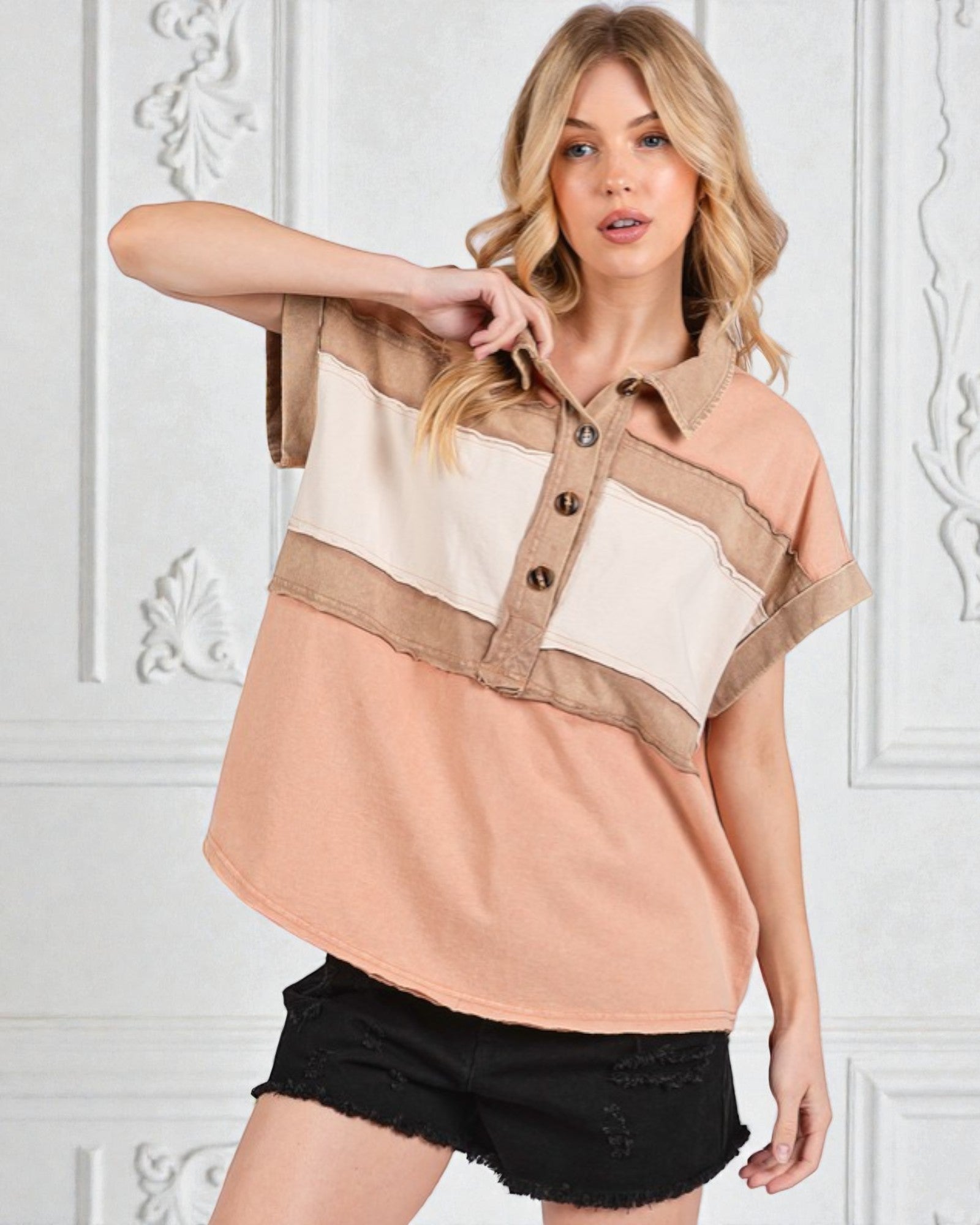 Woman wearing a beige and white color-blocked shirt with black shorts against a white wall.