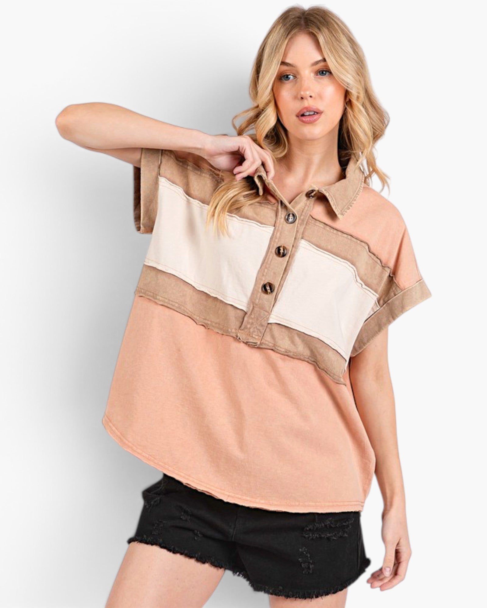 A woman modeling a short-sleeve color-block shirt with a peach, beige, and cream design, paired with black distressed denim shorts, against a light gray studio background.
