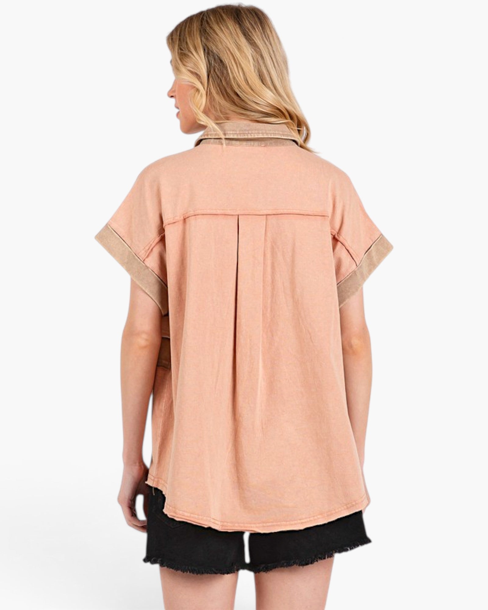 A woman shown from the back wearing a peach-colored short-sleeve button-up shirt with beige contrast trim on the collar and sleeve cuffs, paired with black denim shorts against a white studio background.
