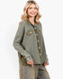 A woman wearing an oversized, washed olive green denim shirt jacket with button-front closure and flap chest pockets, paired with distressed denim pants, against a plain white studio background.
