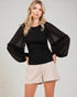 Woman wearing a black top with sheer sleeves and beige shorts on a plain background