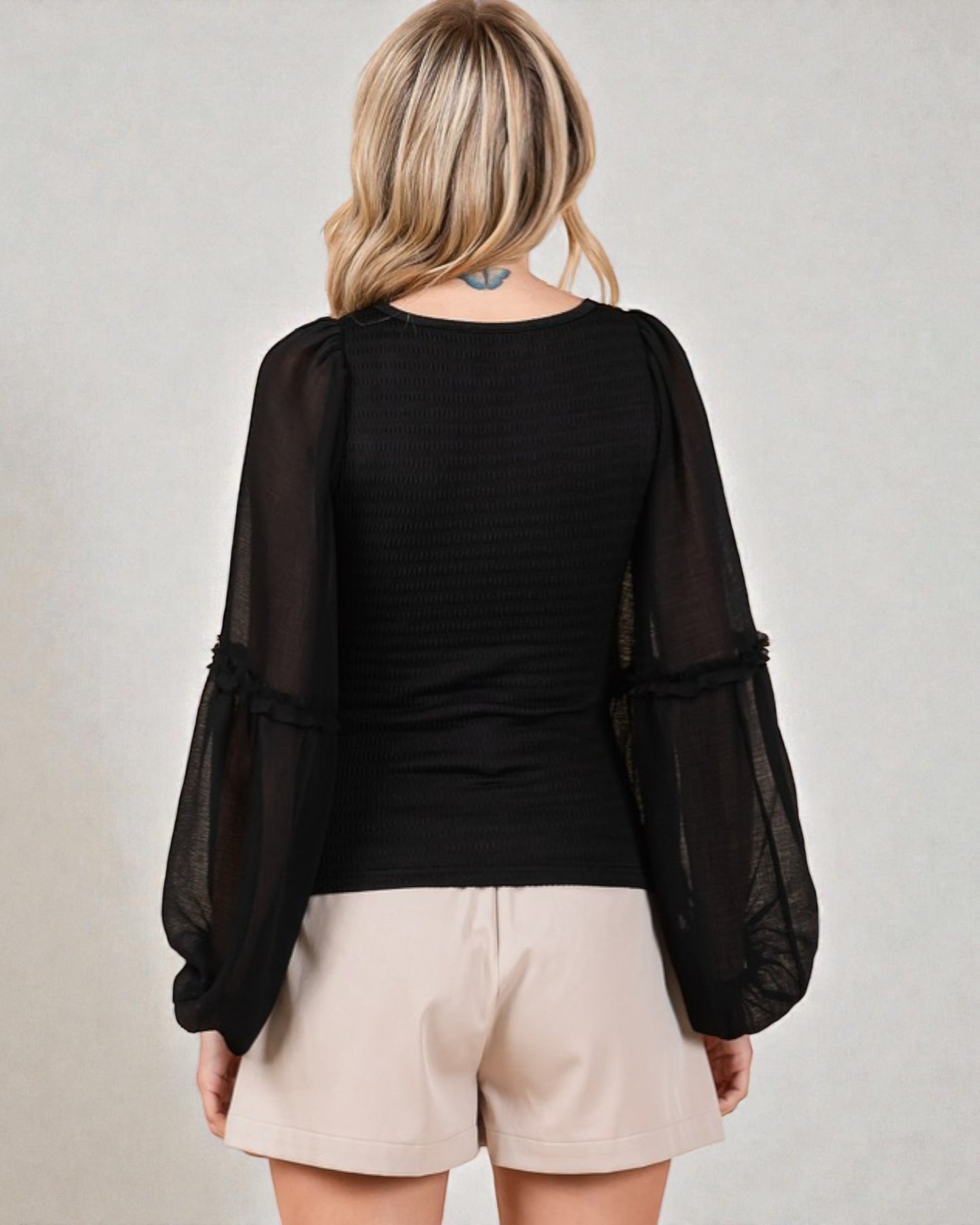 Person wearing a black top with sheer sleeves and beige shorts on a plain background