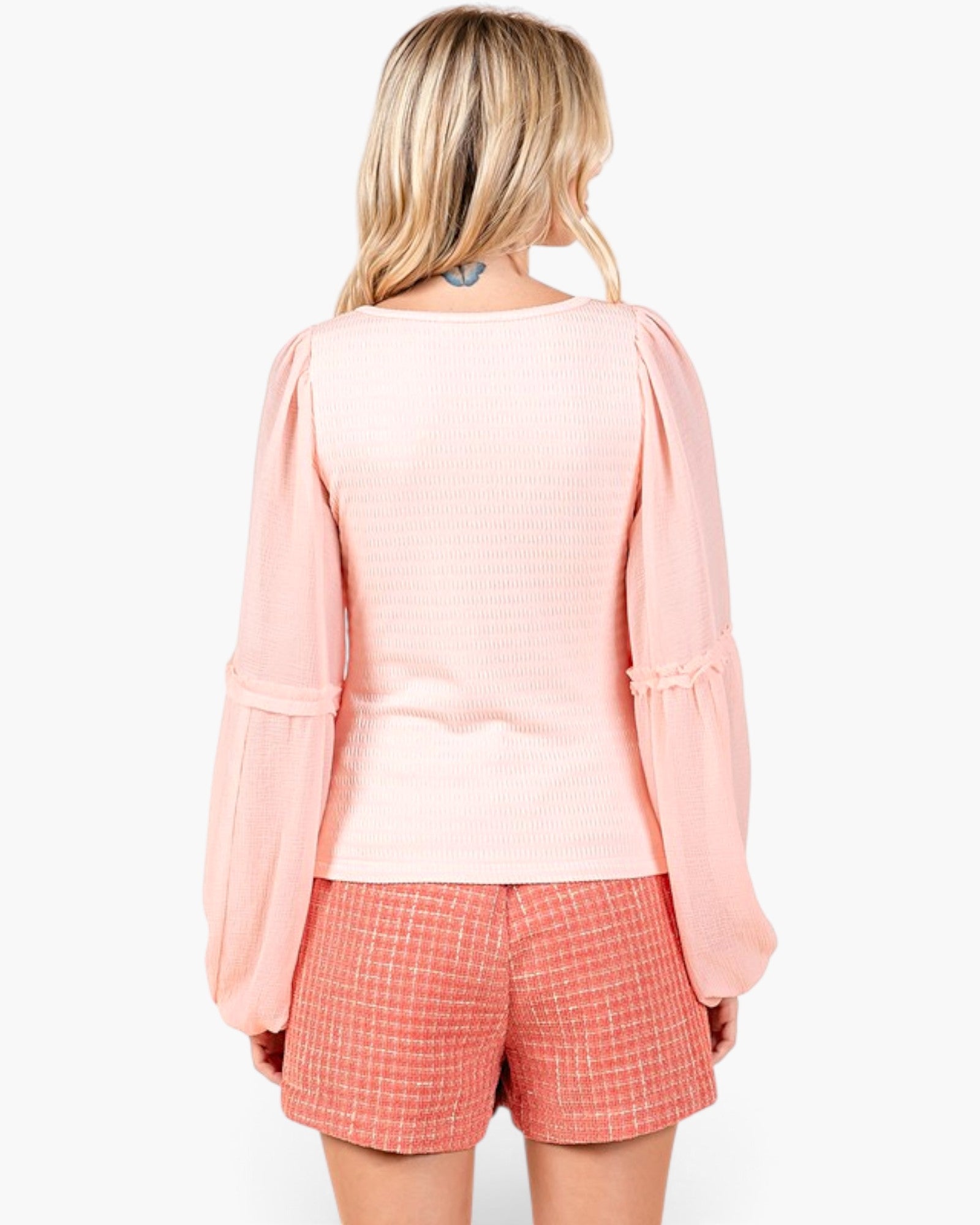 A woman shown from the back wearing a light pink textured long-sleeve top with puffed sleeves and red-and-white checkered shorts against a plain white background.