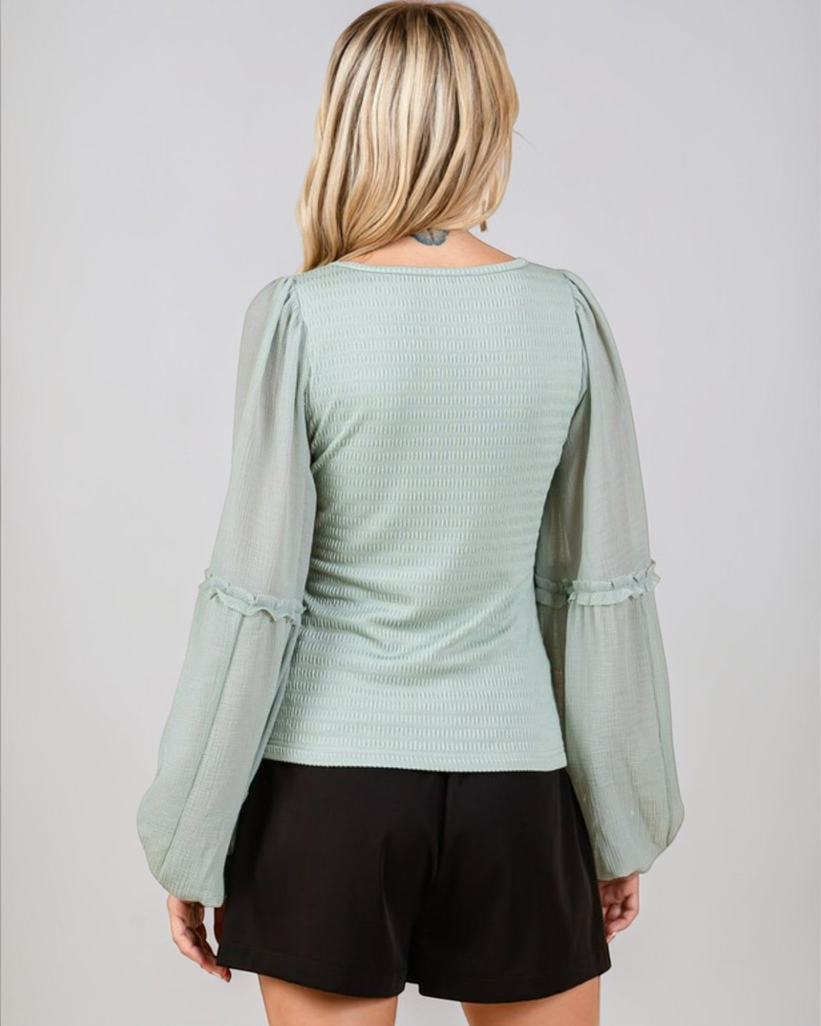 Woman wearing a light green textured top with sheer sleeves against a plain background