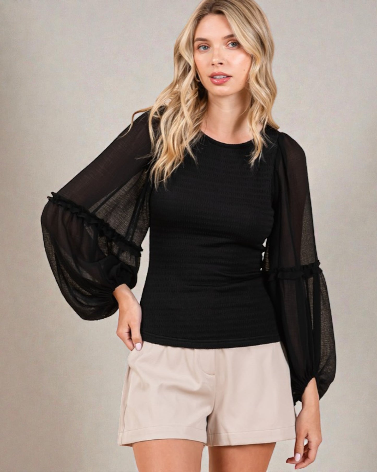 Woman wearing a black top with sheer sleeves and beige shorts on a neutral background