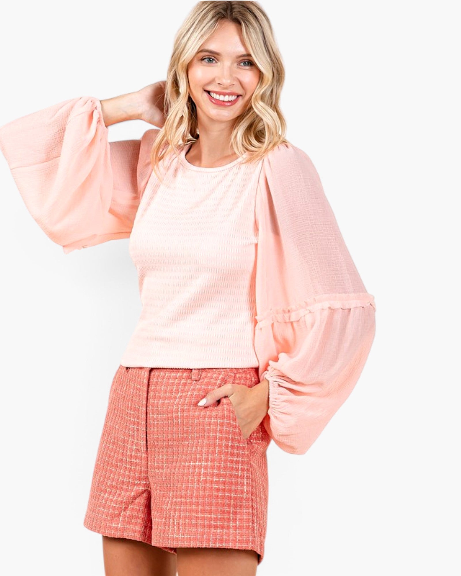 A woman modeling a light pink textured top with dramatic balloon sleeves and matching pink shorts against a plain white background.