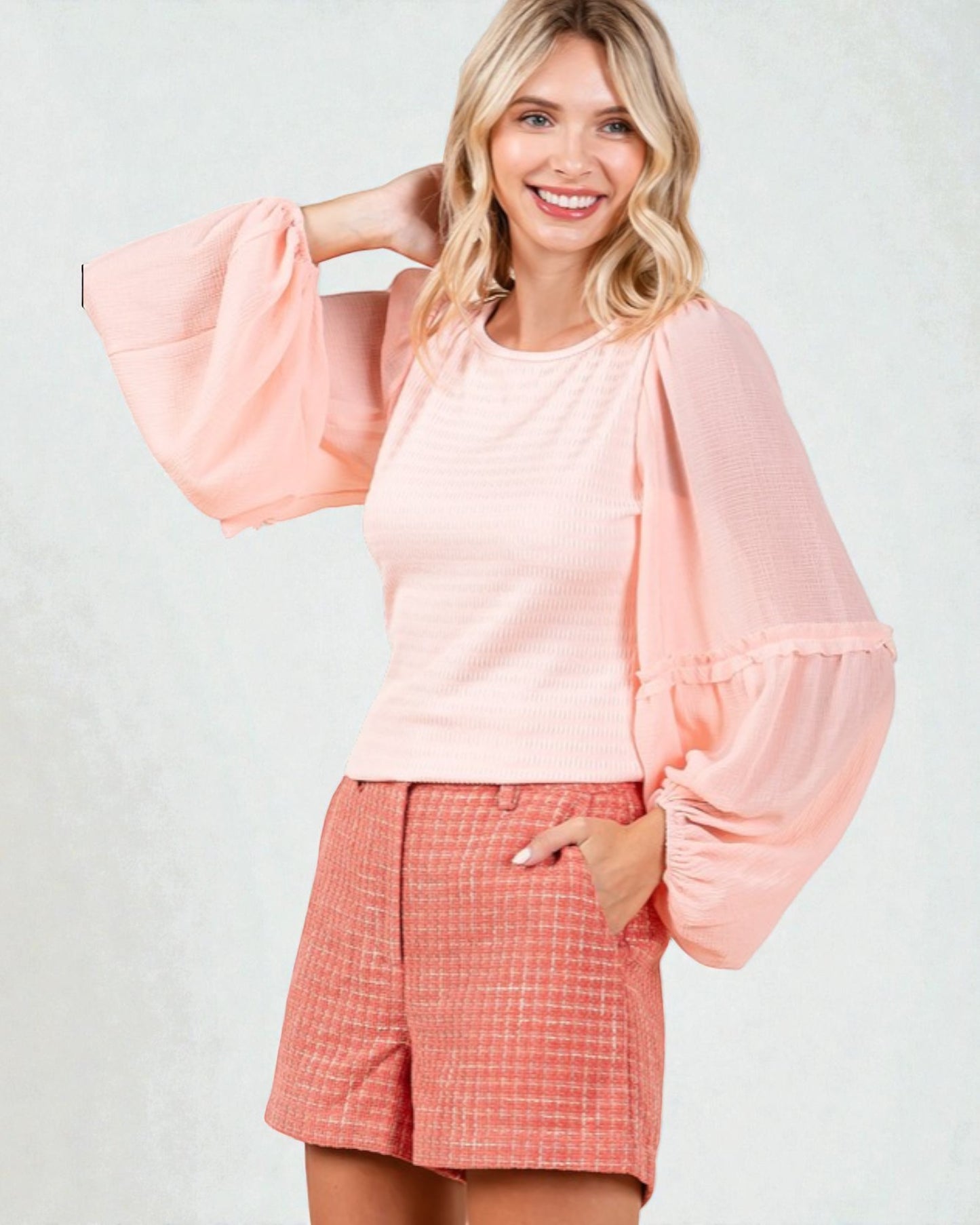 Woman wearing a pink blouse with bell sleeves and coral shorts on a white background