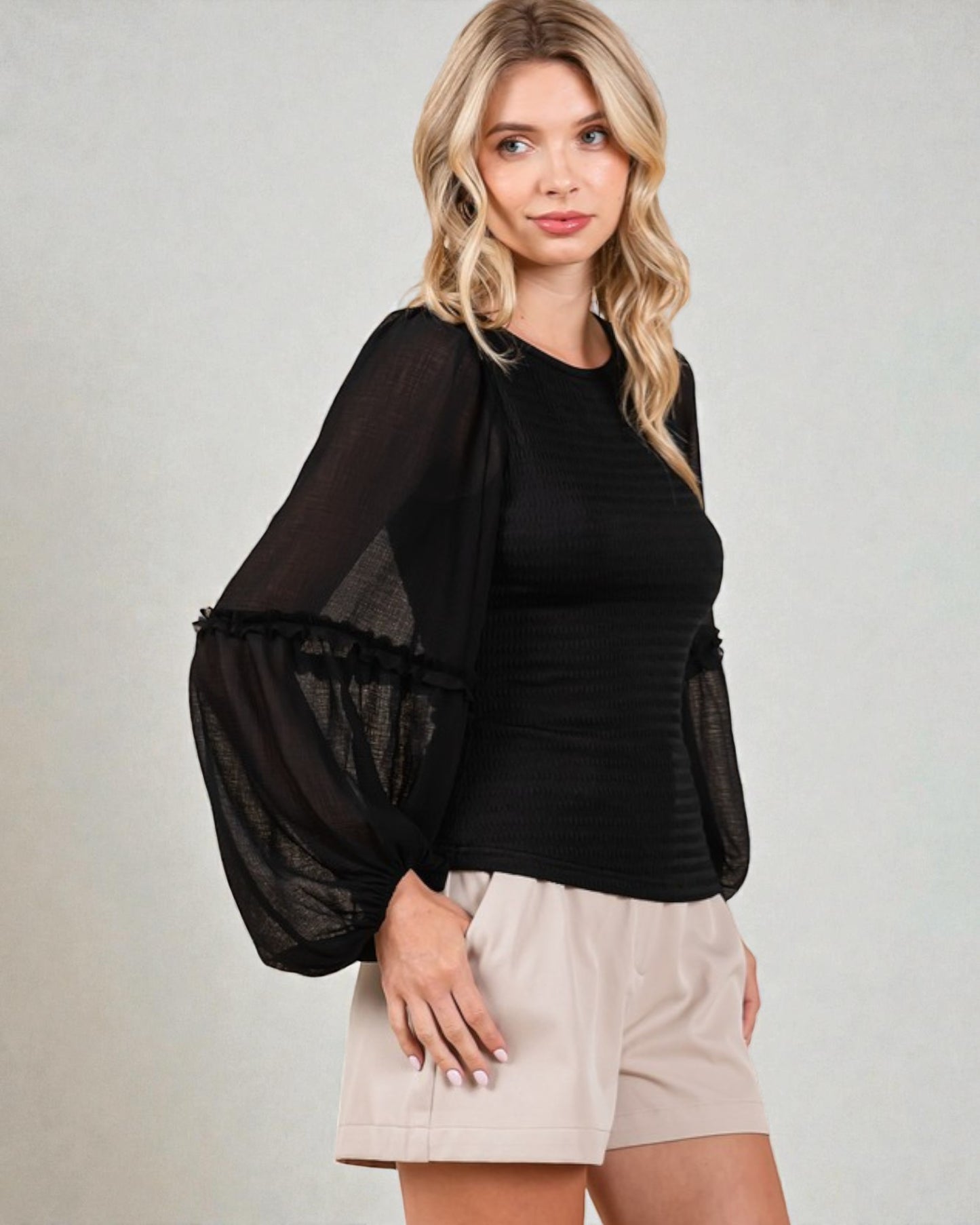 Woman wearing a black top with sheer sleeves and beige shorts on a plain background