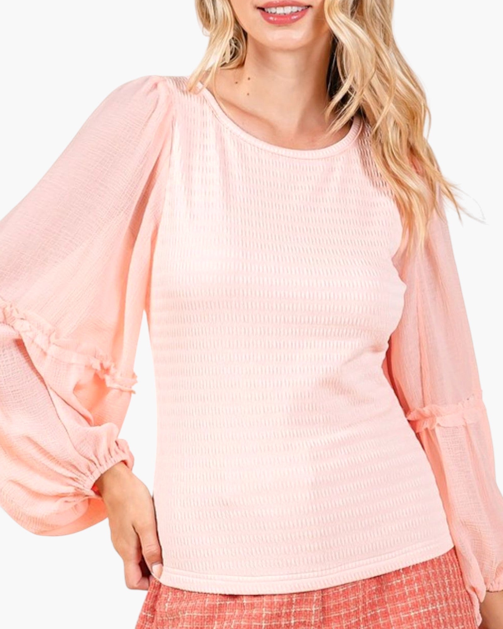 A woman wearing a light pink textured long-sleeve top with voluminous ruffled sleeves and a crew neckline, paired with a pink plaid skirt against a plain white background.
