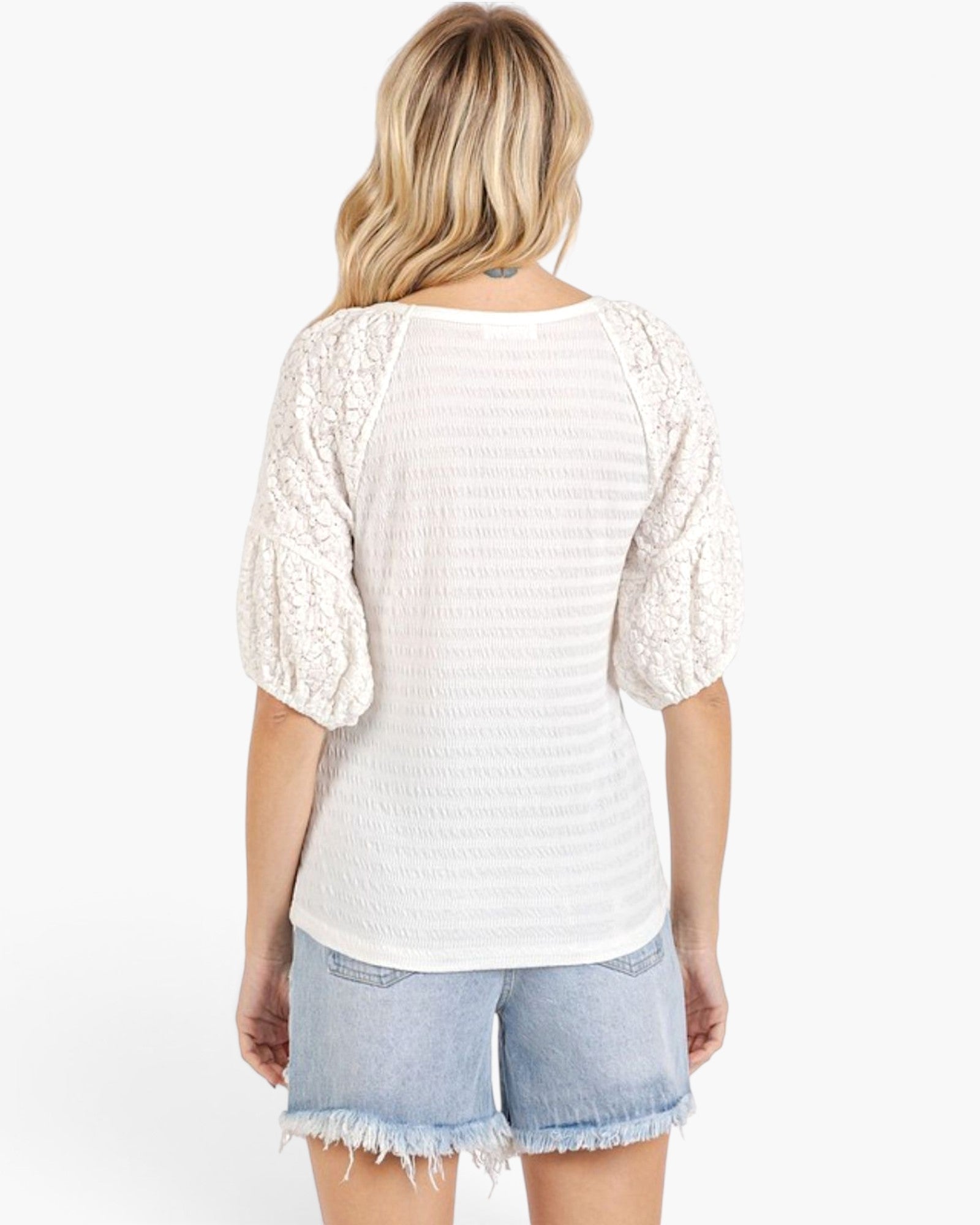 A woman shown from the back wearing a white textured top with lace puff sleeves and light blue denim shorts against a plain white background.