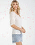 Woman wearing a white lace top and denim shorts against a light background with red abstract shapes.