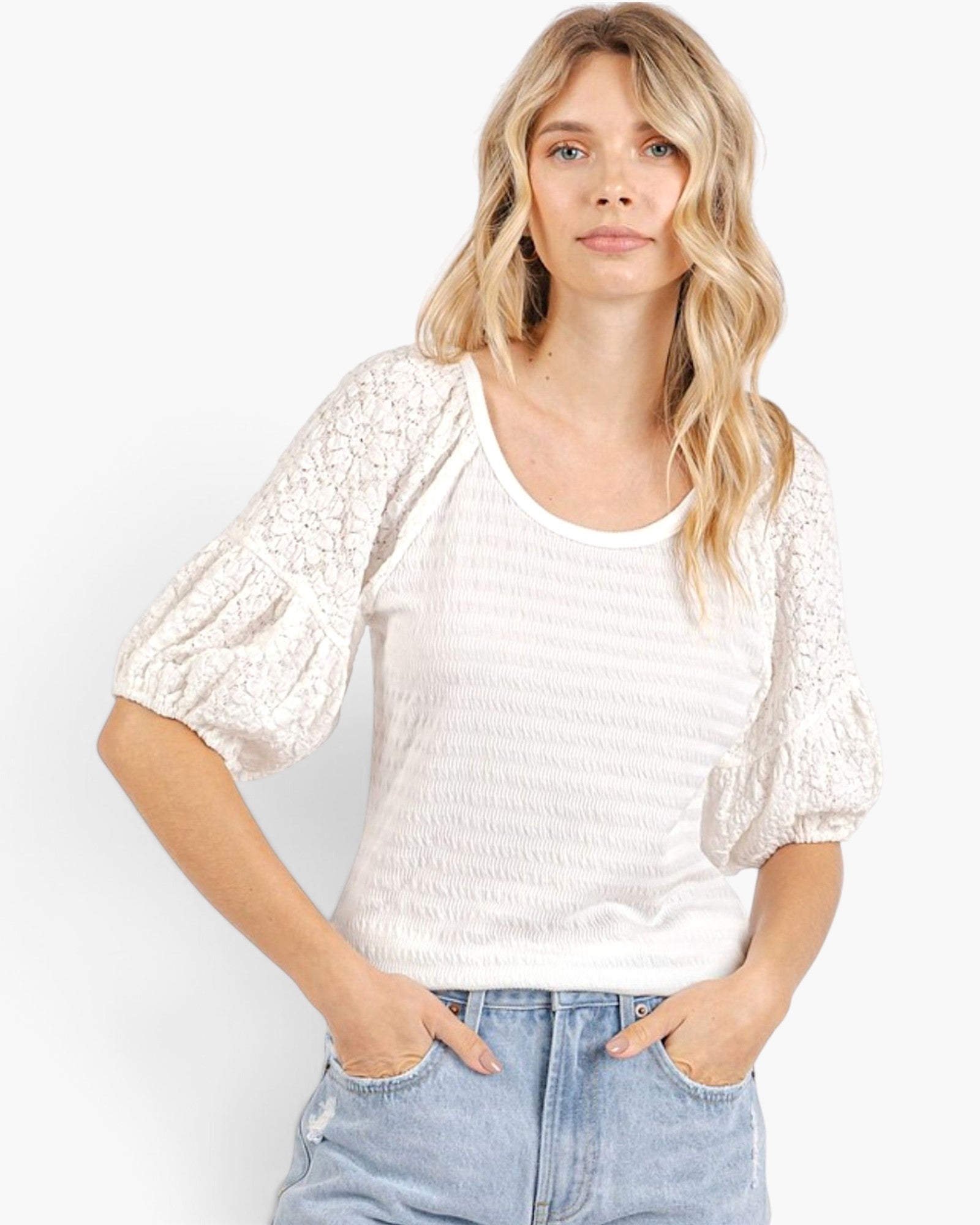A woman wearing a white textured short-sleeve top with sheer lace puff sleeves and light blue jeans, standing against a plain white background.