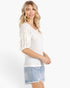 A woman modeling a white textured knit top with three-quarter puff sleeves and a scoop neckline, paired with light blue denim shorts, against a plain white studio background.