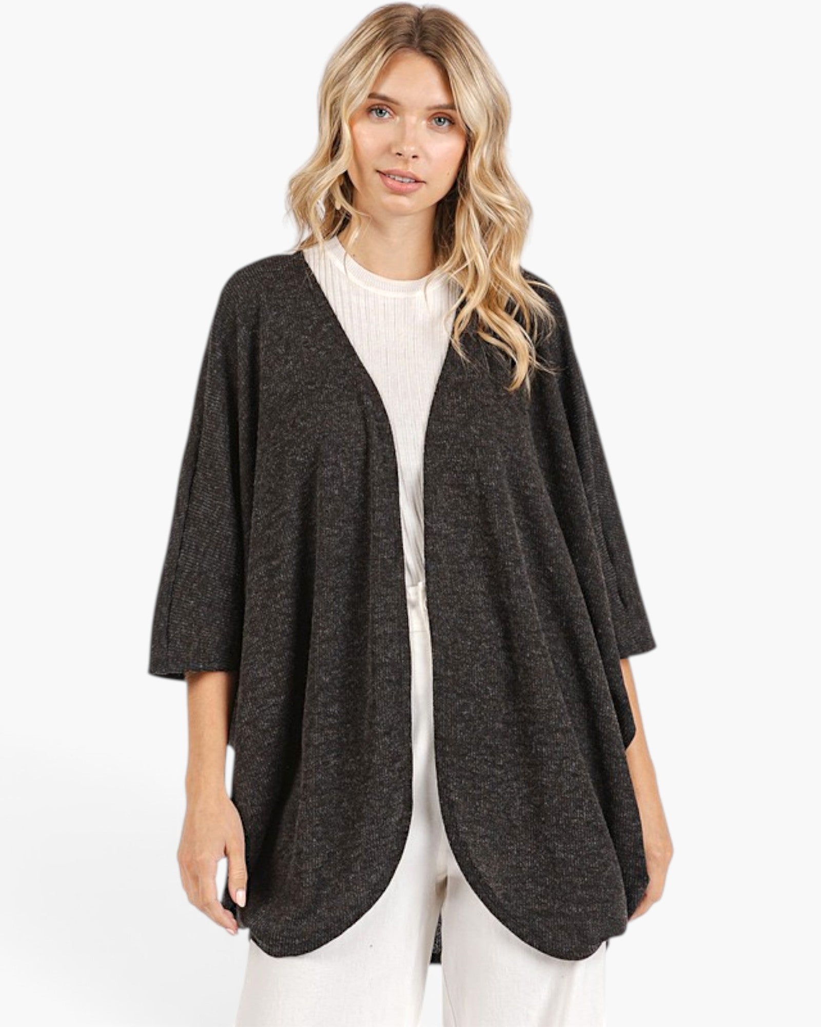 A woman modeling a dark charcoal gray open-front poncho cardigan over a white ribbed top and white pants, set against a plain white studio background.