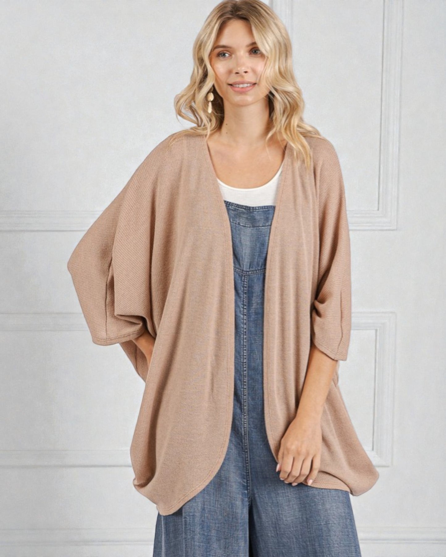 Woman wearing a beige cardigan over a white top and blue denim dress against a white wall.