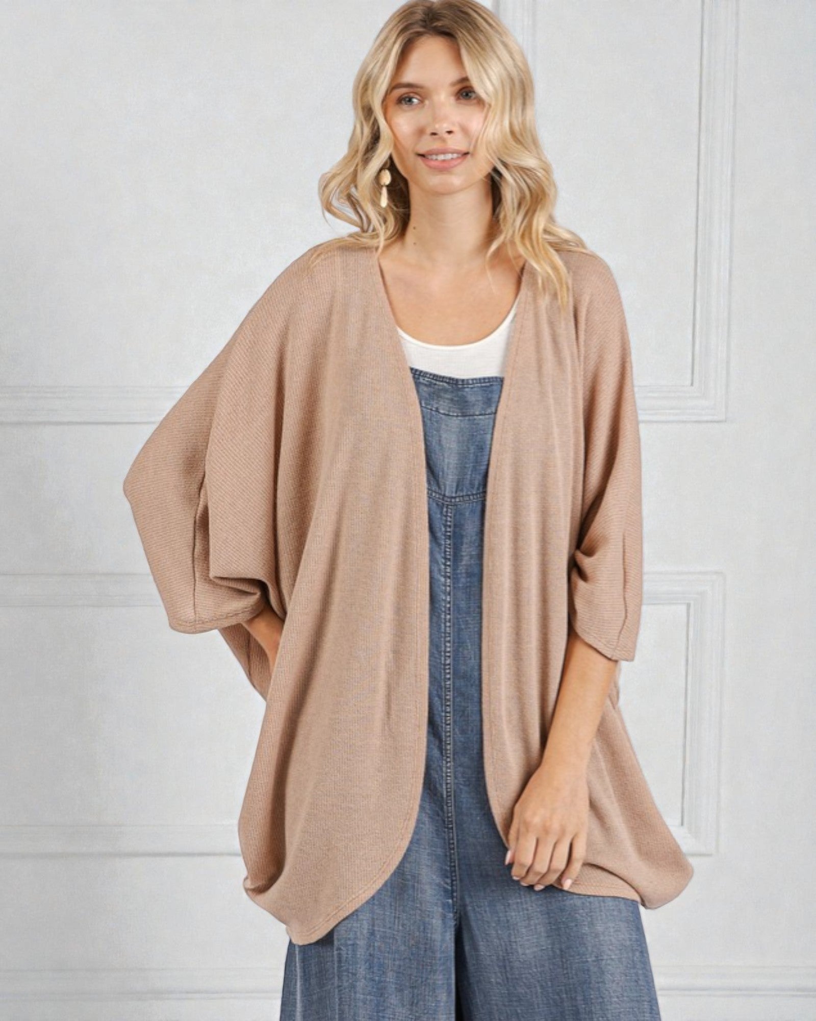 Woman wearing a beige cardigan over a white top and blue denim dress against a white wall.