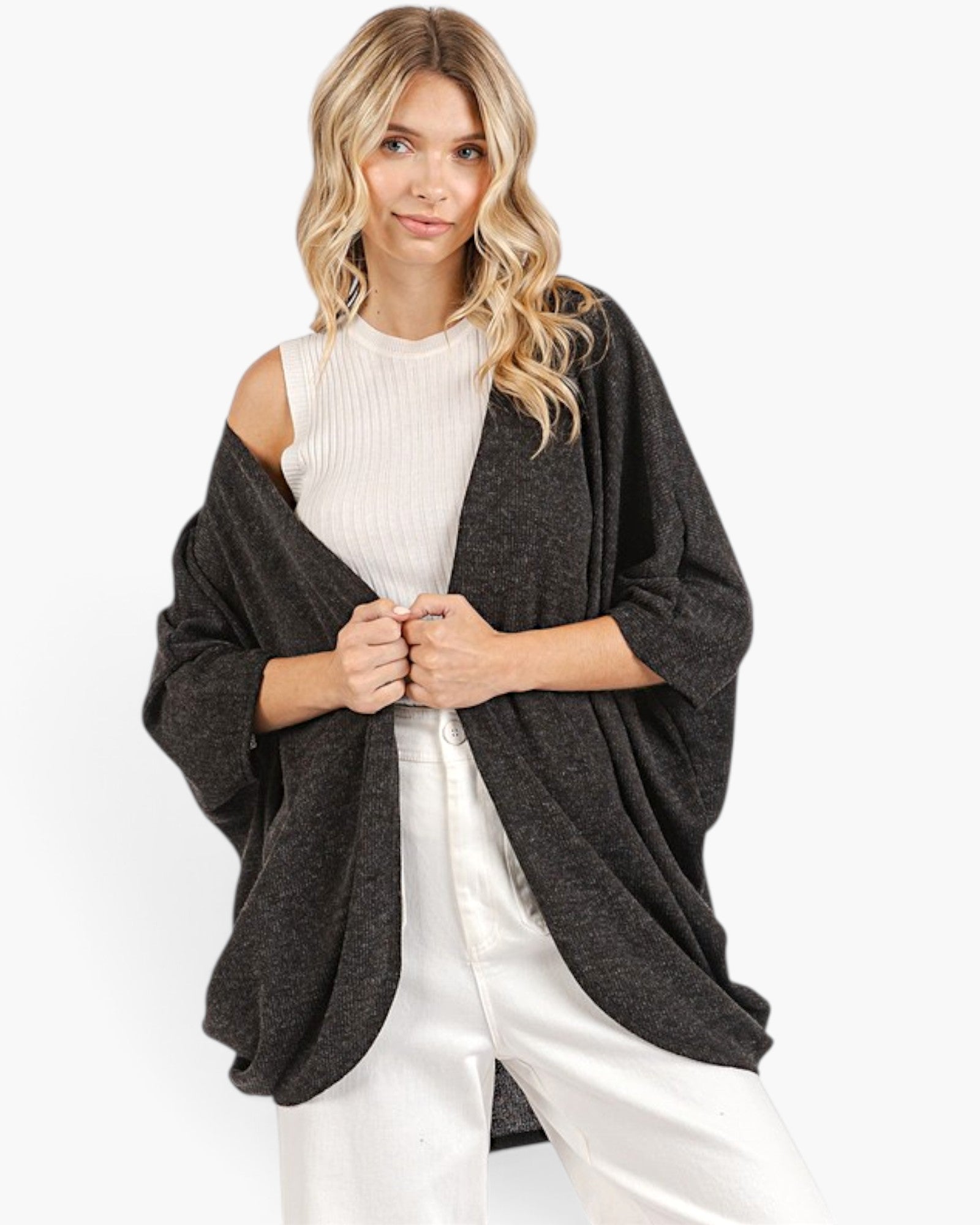 A woman modeling a dark charcoal gray open-front cardigan or poncho layered over a white ribbed cold-shoulder top and white pants, against a plain white studio background.