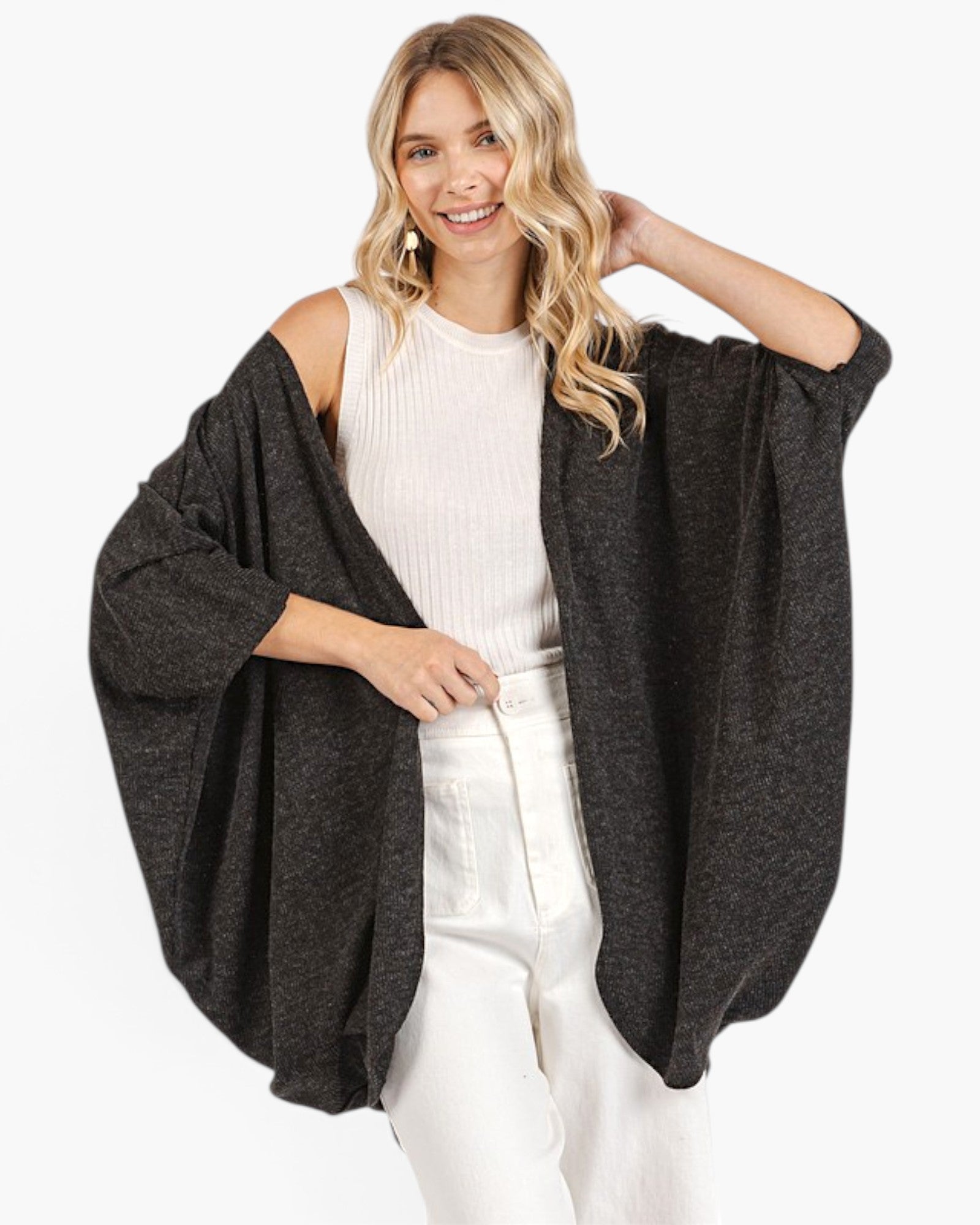 A woman modeling a dark charcoal gray open-front poncho-style cardigan over a white ribbed top and white pants, photographed against a clean white studio background.