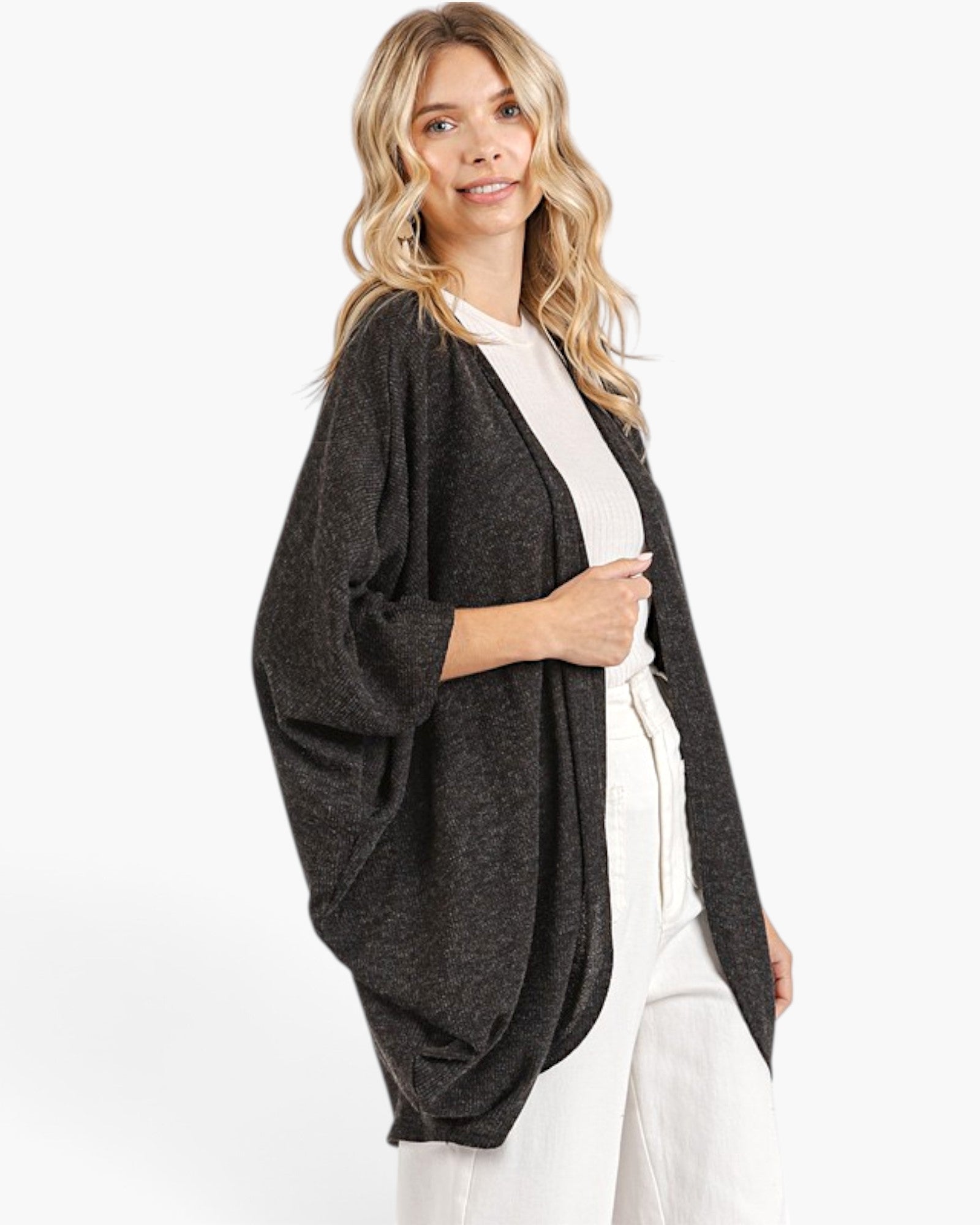A woman modeling a dark gray open-front cardigan with wide three-quarter sleeves, worn over a cream top and white pants against a plain white studio background.