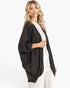 A woman modeling a dark gray open-front cardigan with wide three-quarter sleeves, worn over a cream top and white pants against a plain white studio background.