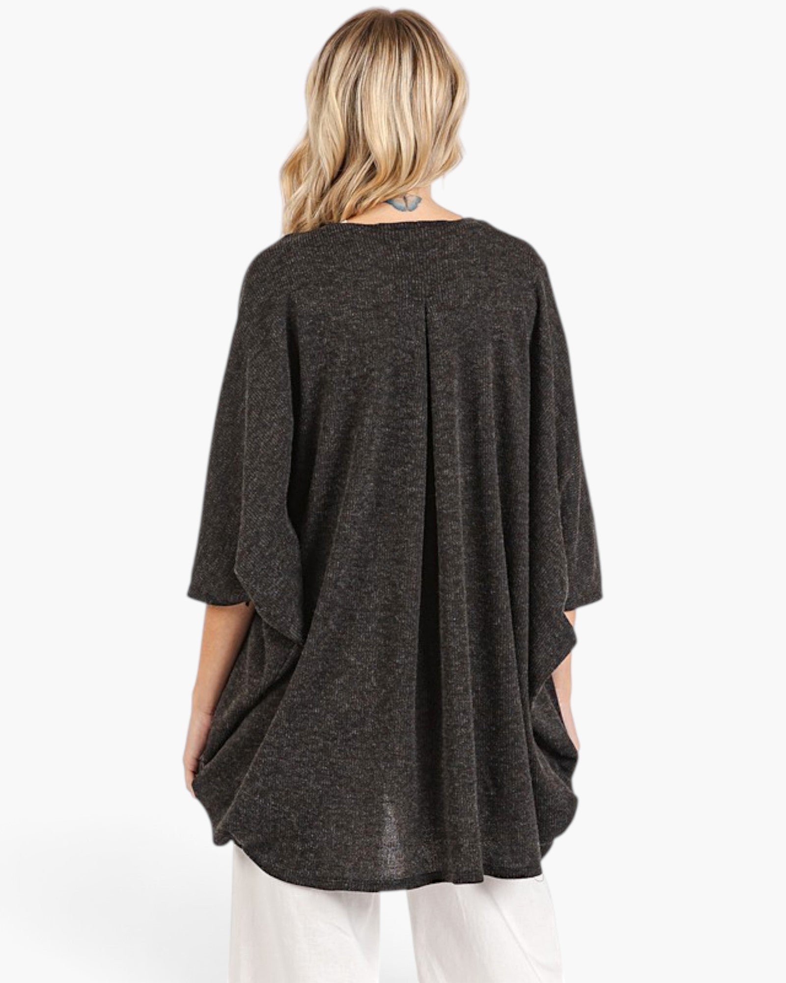 A woman viewed from the back wearing a dark charcoal gray knit poncho-style top with three-quarter sleeves and white pants, against a plain white studio background.