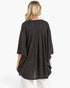 A woman viewed from the back wearing a dark charcoal gray knit poncho-style top with three-quarter sleeves and white pants, against a plain white studio background.