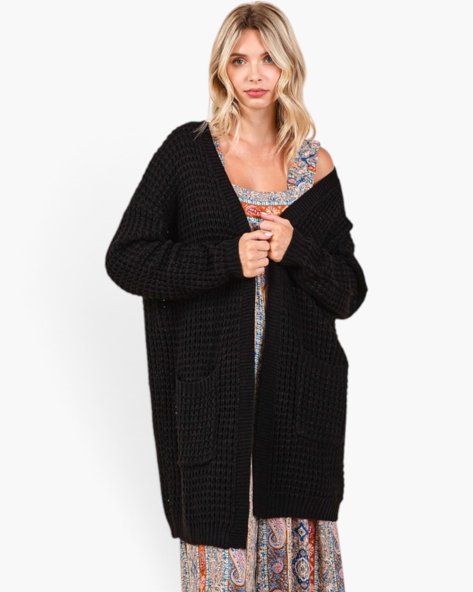 A woman modeling a black open-front waffle-knit cardigan over a patterned dress, photographed against a clean white studio background.