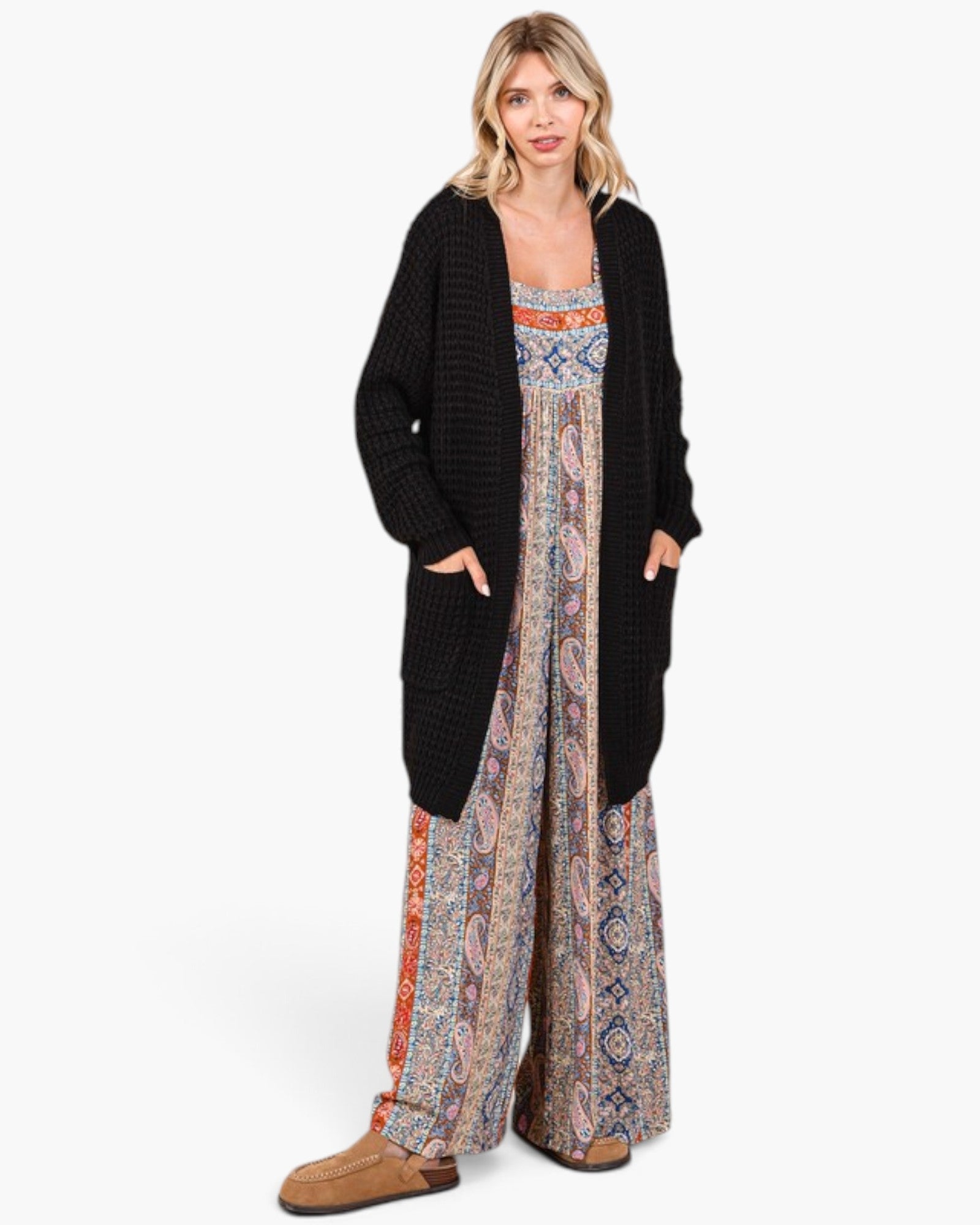 A woman modeling a black textured open-front cardigan over a long-sleeve boho-style jumpsuit with a colorful paisley and geometric pattern, paired with brown suede mules, against a plain white background.