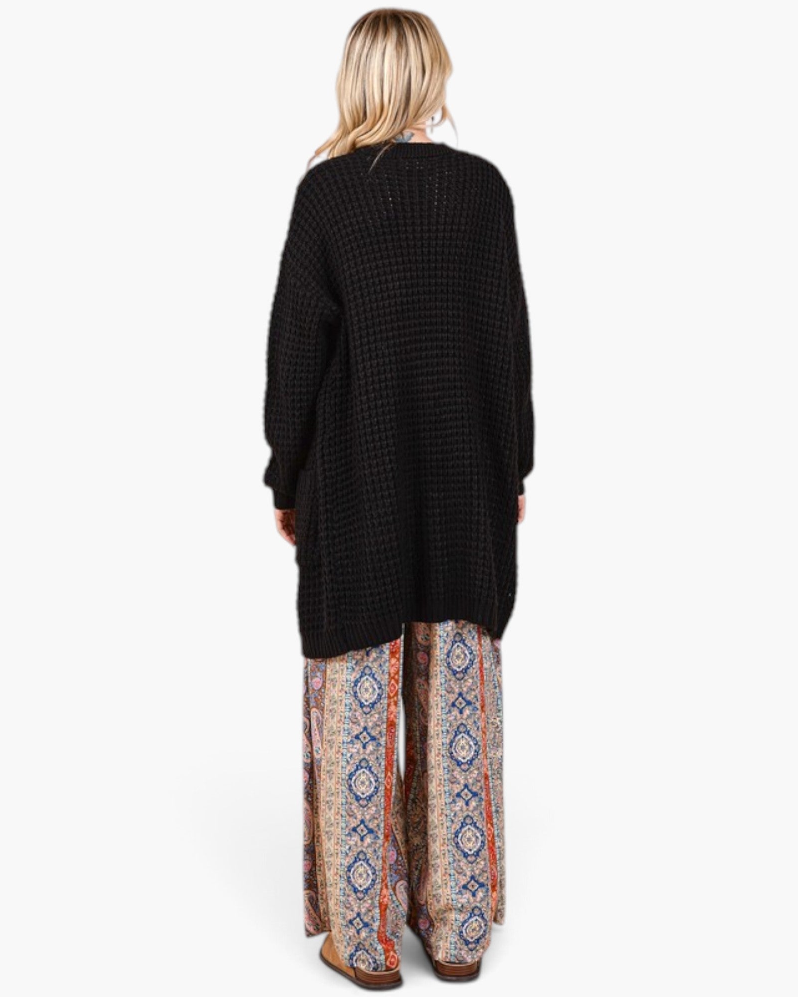 A woman shown from the back wearing a black waffle-knit long cardigan and wide-leg patterned pants with blue, orange, and beige geometric designs, against a plain white background.