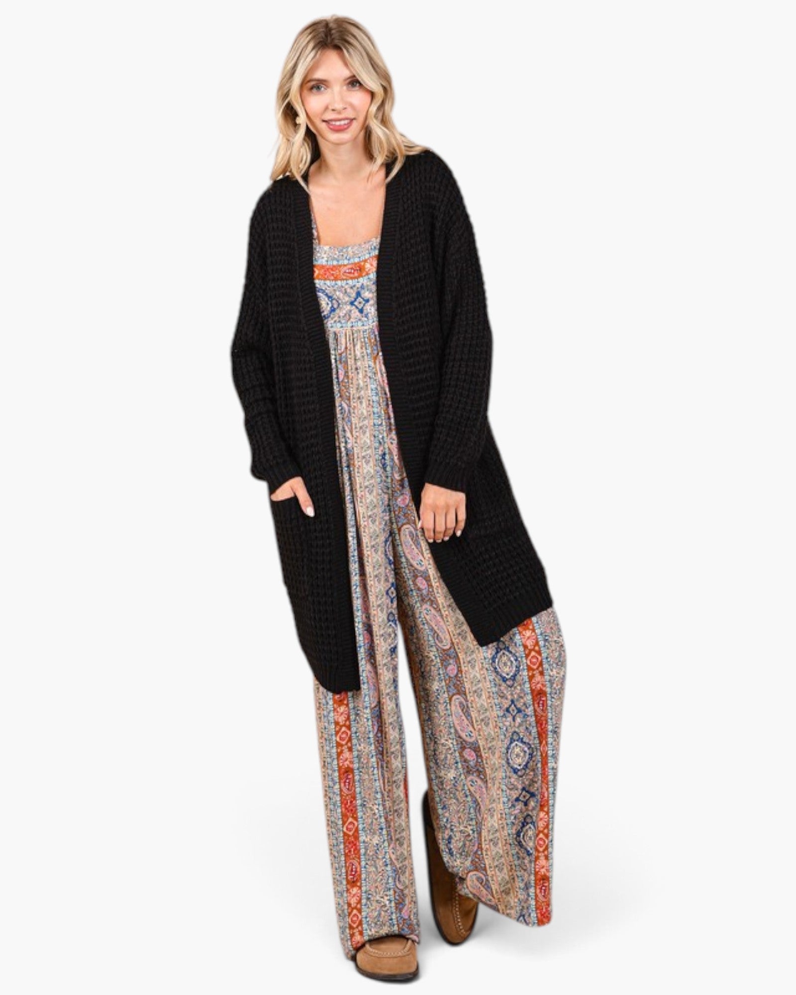 A woman modeling a long black textured cardigan over a patterned bohemian jumpsuit with wide-leg pants and brown boots, set against a plain white studio background.