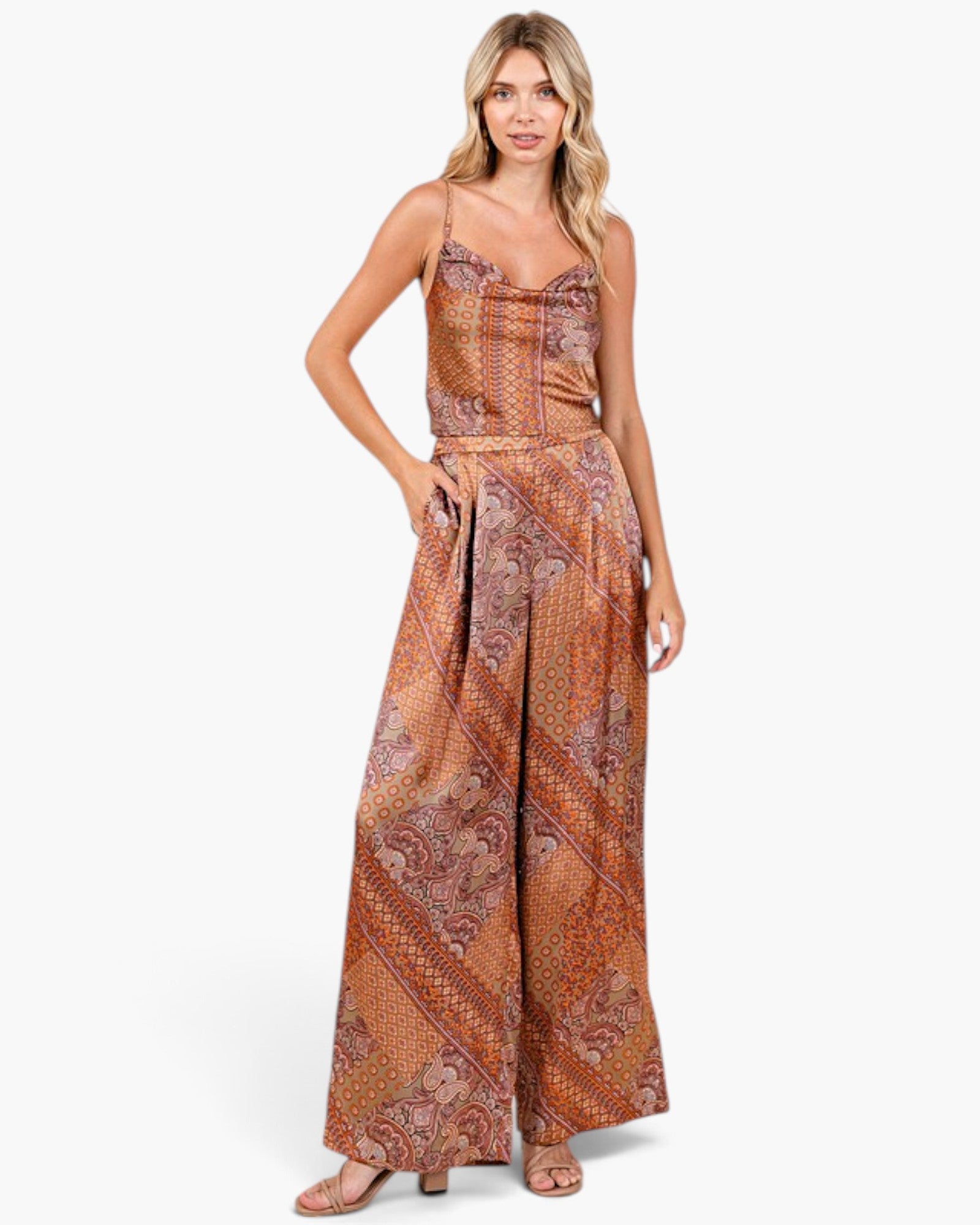 A woman modeling a sleeveless, wide-leg jumpsuit with a brown and rust-orange paisley pattern, photographed against a clean white studio background.