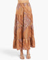 A tiered maxi skirt with an orange, brown, and beige paisley and geometric pattern, shown on a model wearing beige strappy sandals against a light gray background.