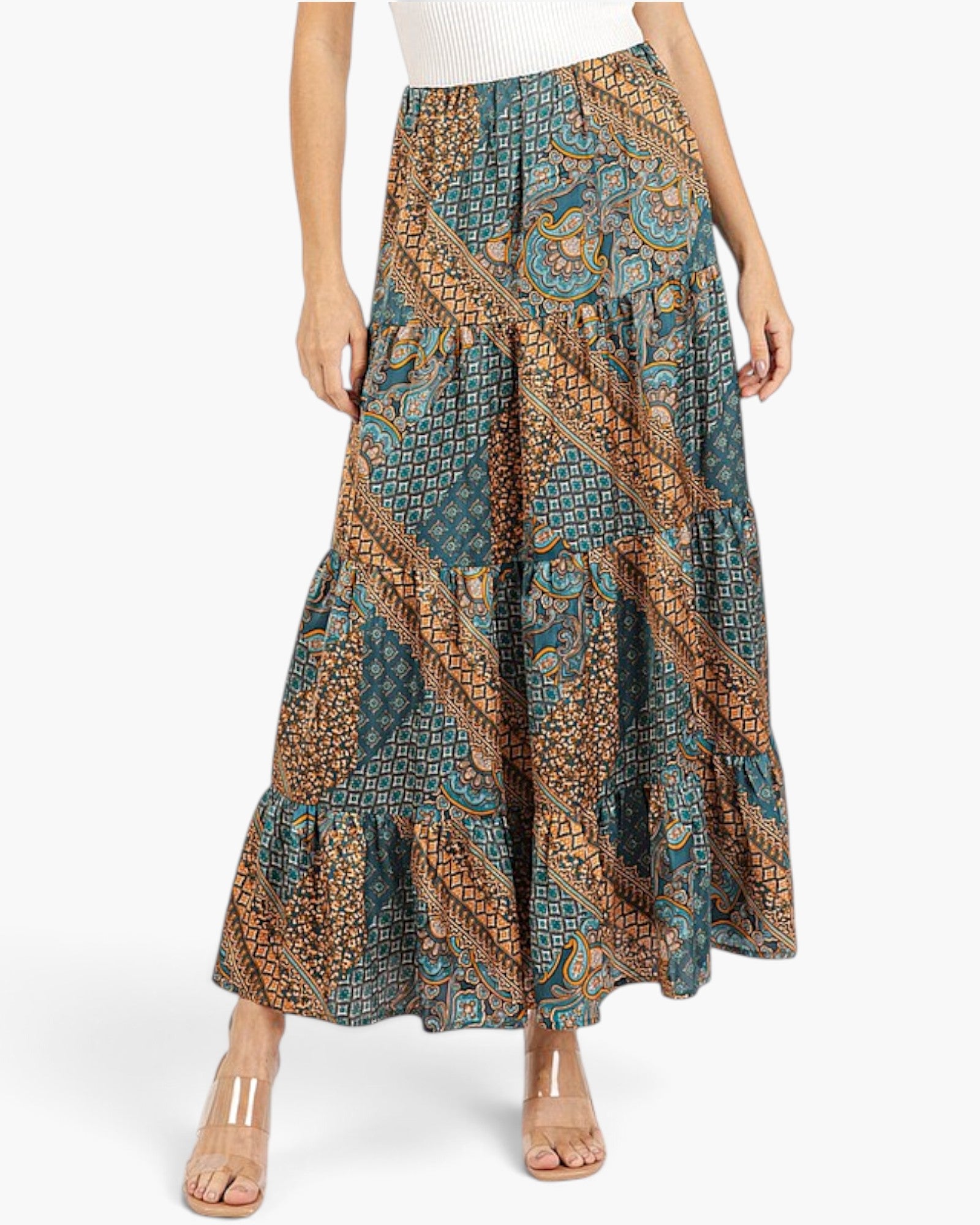 A model wearing a tiered maxi skirt with a complex geometric and paisley print in teal, brown, and gold, paired with clear-strap heels against a plain white background.