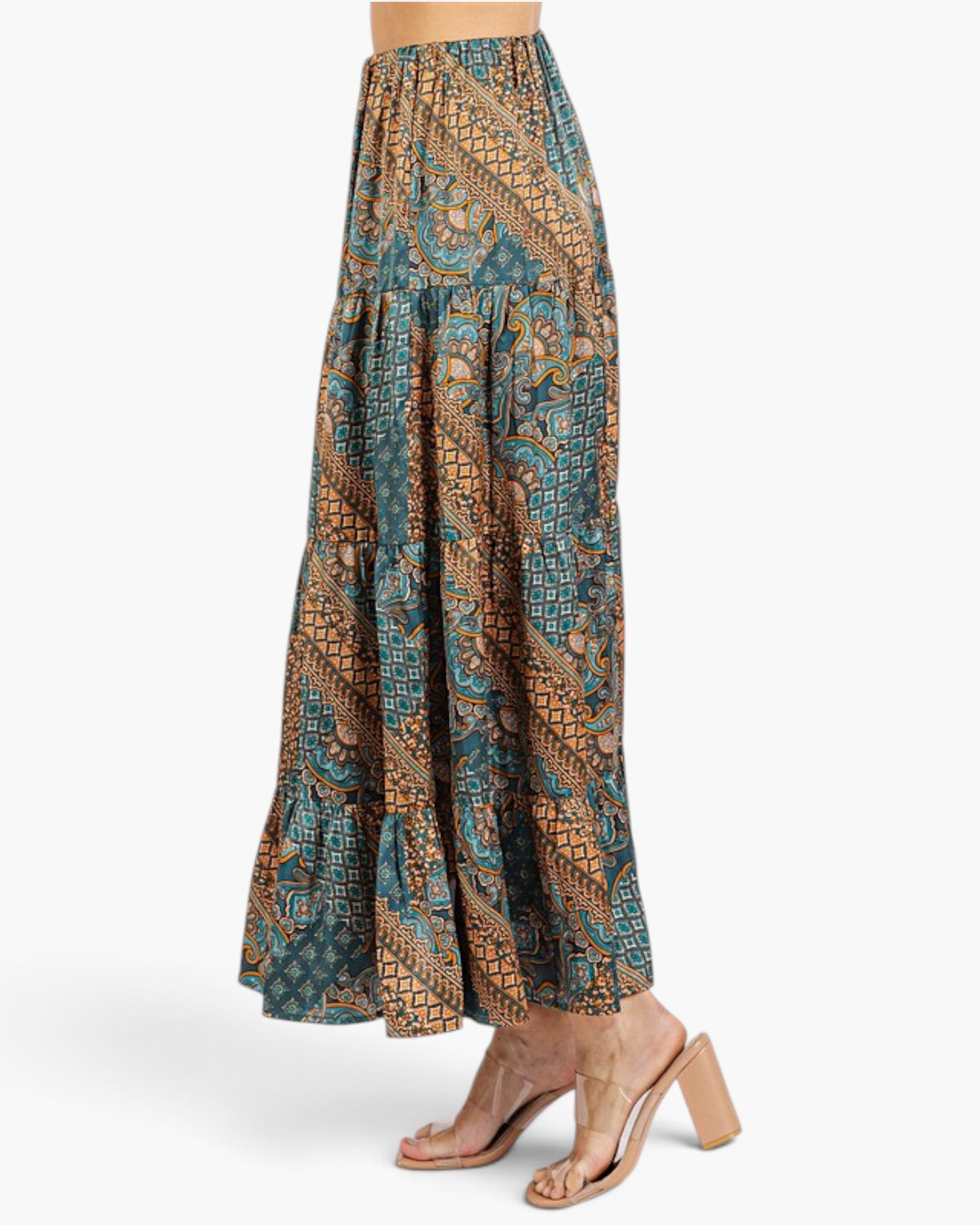 A model is shown from the waist down wearing a tiered maxi skirt with a detailed geometric and paisley pattern in teal, brown, and gold, paired with clear-strap block-heel sandals against a light gray studio background.