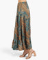 A model is shown from the waist down wearing a tiered maxi skirt with a detailed geometric and paisley pattern in teal, brown, and gold, paired with clear-strap block-heel sandals against a light gray studio background.