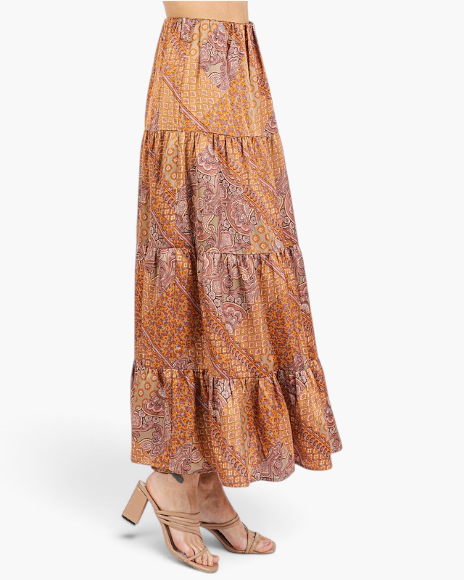 A model wearing a tiered maxi skirt with an intricate paisley and floral pattern in orange, brown, and beige, paired with beige strappy high-heeled sandals against a light gray studio background.
