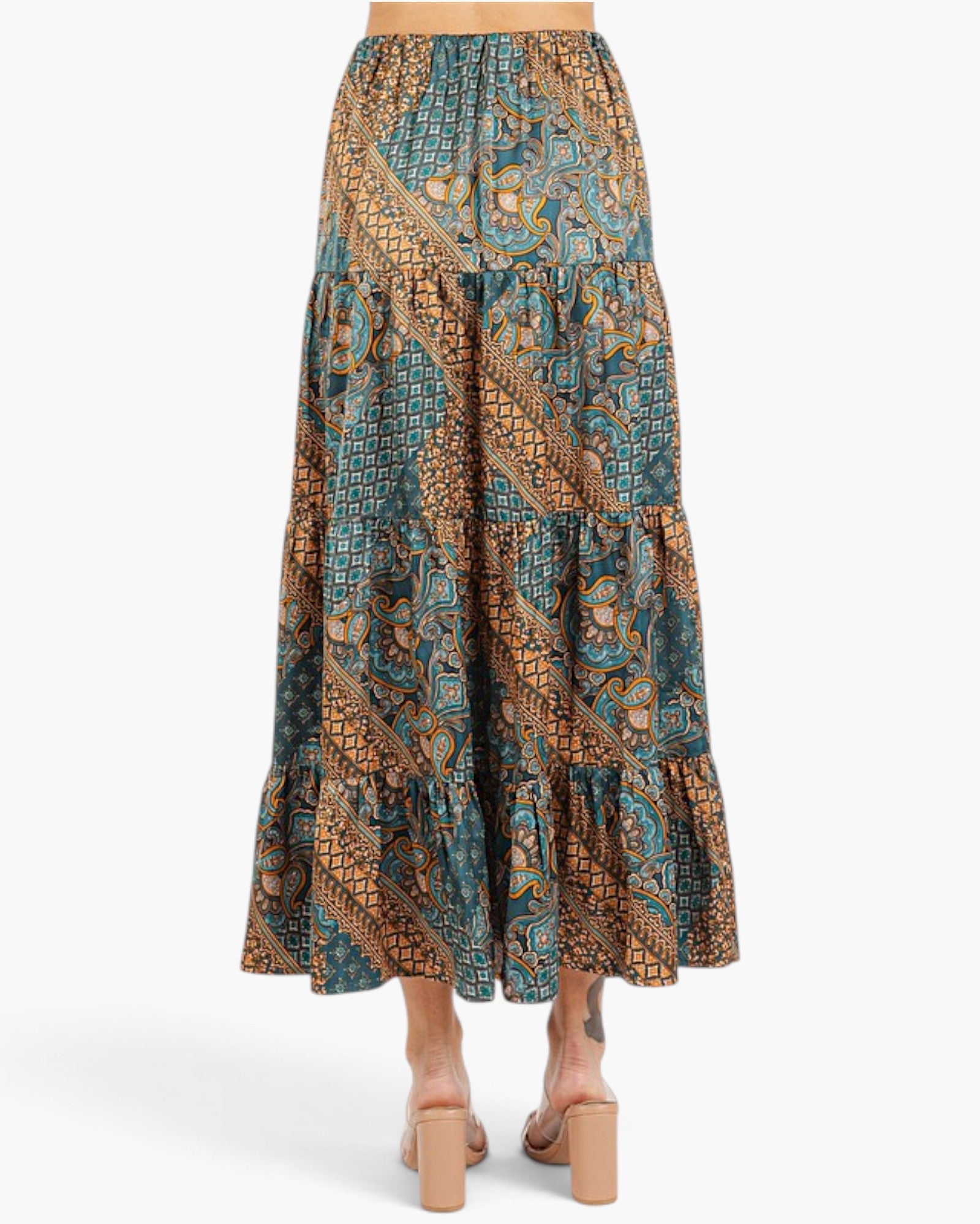 A tiered maxi skirt with a complex batik-style pattern in teal, brown, and gold, shown from the back on a model wearing beige block-heel sandals against a light gray background.